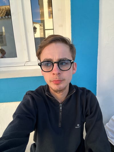 The image is a selfie of a young man wearing black-rimmed glasses and a dark blue zippered sweater. The sweater has a small white logo on the left side of the chest. He appears to be sitting outdoors near a window with white frames and blue walls, possibly in Vila Real de Santo António, Portugal. The window reflects an image of what seems to be buildings or a townscape.