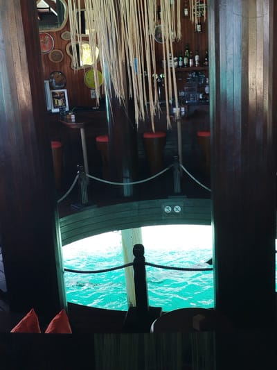 The image depicts the interior of a rustic bar with wooden paneling and decorative elements, overlooking a vibrant turquoise ocean. In the foreground, two bright orange triangular objects, possibly life vests, are visible on a dark surface. A rope barrier separates the bar area from an open section that looks out onto the water. Through the opening, the clear, shimmering ocean is visible, with gentle waves and what appears to be the bottom of a boat or structure submerged beneath the surface. In the background, the bar area features shelves stocked with bottles of alcohol, bar stools with orange seats, and various decorative items on the wooden walls, including mirrors and circular plates. Hanging strands of what appear to be dried reeds or bamboo further adorn the space, adding to the tropical ambiance. The overall impression is a cozy, nautical-themed bar in a scenic Maldivian location. Based on the context of Gulhi Falhu, Maldives, this scene likely represents a bar or restaurant situated on a boat or a structure built over the water, offering patrons a direct view of the ocean.