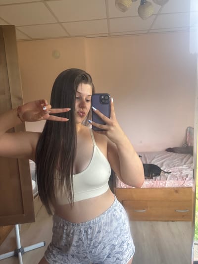 The media file is a vertical, full-body mirror selfie taken by a young woman. She has long, dark hair that is covering part of her face. She is wearing a white ribbed tank top and patterned shorts. The woman is holding a dark-colored smartphone in her right hand, which she is using to take the picture. Her left hand is raised, forming a peace sign. She is looking at the camera and appears to be pouting slightly.
The setting is an indoor space, likely a bedroom. In the background, reflected in the mirror, is a bed with pink and white bedding, and a black dog lying on it. There is also a wooden dresser with two drawers and a window with blinds. The lighting suggests it is daytime. The room appears to be a domestic setting. The overall mood is casual and playful. There is no visible text in the image.
