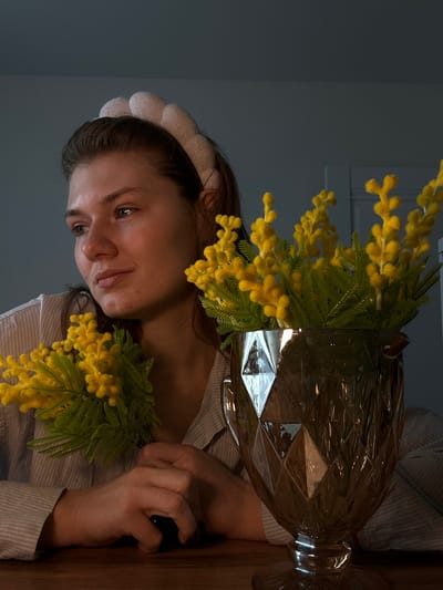 An indoor scene in Nikol'skii, Russia, depicts a female subject seated at a dark wooden table. She wears a light-colored, padded headband and a light-colored, vertically striped shirt. Her hands are clasped on the table surface, and she holds a small cluster of yellow, mimosa-like flowers with green foliage in her left hand. Her head is turned slightly to the left, and her gaze is directed towards the left of the frame. To the right of the subject, a larger arrangement of identical yellow flowers is presented in a clear, faceted glass vase with a footed base. The background consists of a dimly lit, light-colored wall, with what appears to be a door frame visible on the right side.