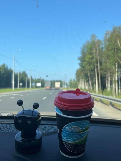 A black dashboard is visible in the foreground, with a black robotic figurine with two antennae and heart-shaped eyes positioned on the left. To the right of the figurine, a black disposable coffee cup with a red lid is placed. The cup has a scenic design featuring a body of water and green hills.

The background shows a highway scene through a windshield that has some dust and smudges on it. The highway has white lane markings and is bordered by a barrier on the right. Trees line the right side of the highway, and power poles are visible on the left. Several trucks are driving on the highway in the distance. The sky is clear and blue.

This image was taken while stationary in a vehicle on a highway, likely in or near Il'yata, Russia, based on context provided. The scene depicts a moment of rest or observation during travel, with personal items placed on the dashboard.