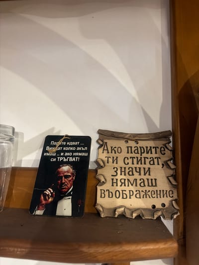 The image shows two decorative plaques on a shelf. The left plaque features a black background with a portrait of a man in a tuxedo, likely Marlon Brando as Don Corleone from "The Godfather." Text in white, Cyrillic script is above the portrait. The right plaque is made of a lighter material, resembling wood, with a wavy, scroll-like design and Cyrillic text carved into it. Both plaques are hung by twine. The shelf appears to be made of dark wood, and a portion of a glass jar is visible to the left of the plaques. The background is a plain, light-colored wall with some shadow play. The location is identified as being in Beguntsi, Bulgaria.