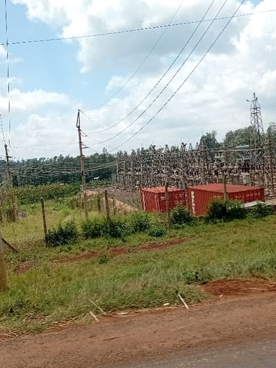 The image displays an outdoor scene of an electrical substation in Mururi, Kenya, under a partially cloudy blue sky. In the immediate foreground, a reddish-brown dirt road or shoulder is visible, transitioning into a grassy verge. A chain-link fence, supported by wooden posts, separates this foreground area from the main substation property. Within the fenced perimeter, two red standard shipping containers, one clearly labeled "MAERSK," are positioned on the ground. The electrical substation occupies the mid-to-background, characterized by a dense complex of grey metal structures, insulators, transformers, and various electrical components. Numerous overhead power lines extend across the frame, supported by utility poles, with one prominent pole in the mid-left. A tall, latticed metal transmission tower stands on the right side of the substation compound. A line of dense green trees forms the distant horizon behind the substation. No individuals are present in the scene.
