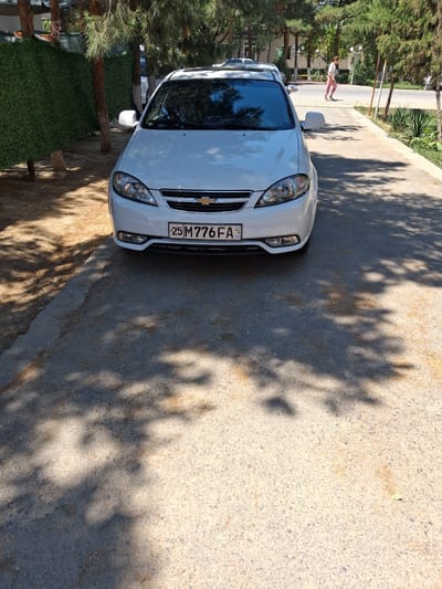 A white Chevrolet sedan is parked on a paved road in Kahramon, Uzbekistan, under the shade of trees casting dappled shadows. The car's license plate is visible, displaying "25 M 776 FA" with a Uzbek flag emblem. To the left of the car is a dense green hedge. In the background, a woman in pink pants and a white top is walking on a sidewalk. Other trees and buildings are visible in the distance.