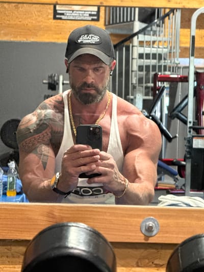 An image, originally rotated 90 degrees counter-clockwise, captures a muscular man taking a mirror selfie in a gym. The man, depicted from the chest up, has a serious or focused expression, looking directly into the smartphone he holds. He wears a dark baseball cap with the mirrored text "OLD DUDE" visible on its front. He also has a beard and mustache.
He is wearing a white tank top, and part of the mirrored text "COMPOUND" is visible on the chest area. His physique is well-developed, with prominent musculature and visible veins, particularly on his shoulders and arms. His left arm (appearing on the right in the image due to the selfie perspective) is covered in extensive black and red tattoos. Accessories include a gold chain around his neck, a gold bracelet on his left wrist, and a watch with a yellow face on his right wrist. He holds a black smartphone, which appears to be an iPhone with a triple camera setup, in both hands to capture his reflection.
The background, reflected in the mirror, shows various gym equipment such as weight racks and benches. A wooden pillar or wall section is visible on the right, featuring a round metal bolt. On the far left, a black sign with white, illegible text is visible. Below the mirror, two plastic water bottles are present. The setting is an indoor gym environment, potentially located in Jemelle, Belgium.
