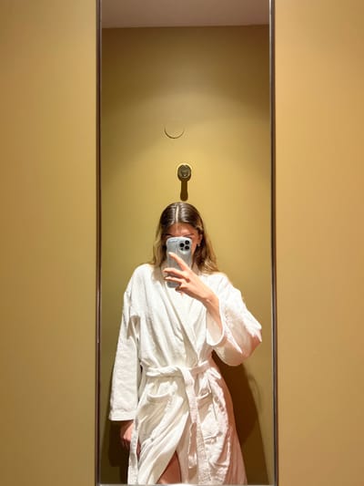 The image shows a woman in a white bathrobe standing in front of a mirror. She is holding a smartphone in front of her face, covering her eyes and mouth. Her left arm is extended, and her right arm is slightly bent, holding the smartphone. The woman has long, dark brown hair and is wearing a white bathrobe with a sash. The walls of the room are a light beige, and the bathroom is lit by artificial light. The bathroom appears to be located in a hotel in Val-d'Isère, France. There are no other people in the image.