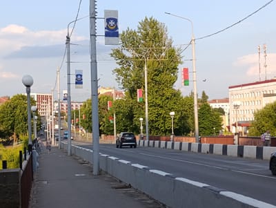 The image depicts a street scene on a bridge in Orsha, Belarus. The time of day appears to be late afternoon or early evening, with a clear sky and soft lighting. The bridge is lined with streetlights and decorated with several flags, some of which appear to be the national flag of Belarus.
On the sidewalk to the left, two women are walking. A black car is driving on the bridge, its rear visible to the viewer. In the background, there are buildings, trees, and other vehicles, suggesting an urban environment. The mood of the scene is calm and ordinary, depicting a typical day in the city. There are no discernible emotions or dramatic events taking place.
The flags displayed are a notable detail, indicating a potential public event or commemoration. The architecture of the buildings and the design of the street furniture provide clues about the specific location within Orsha. The overall impression is that of a functional and well-maintained urban infrastructure.
