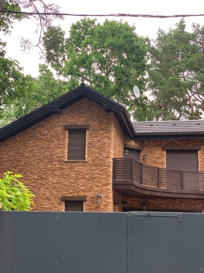 The image shows a two-story house with a stone veneer facade in Pobierowo, Poland. The house has a dark-colored roof and dark-colored roll-down shutters on its windows. There is a wooden balcony with a slatted railing on the second floor. Green trees surround the house, and a dark grey fence is visible in the foreground. A satellite dish is mounted on the roof.
