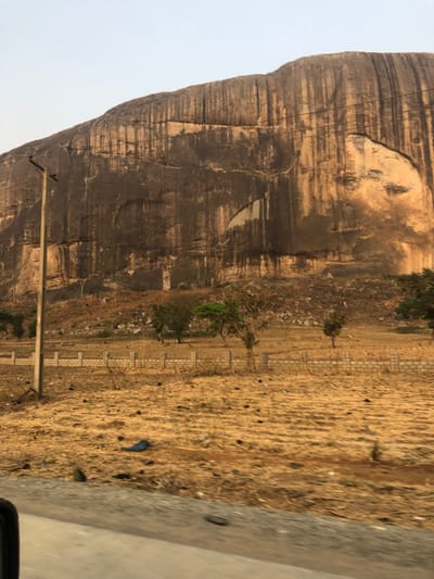 The image displays a vast outdoor landscape dominated by a massive rock formation under a bright, overcast sky. The photograph appears to have been taken with the camera rotated 90 degrees clockwise, making the rock formation appear to stretch horizontally across the left side of the frame, and the ground along the right.
Upon correcting the orientation mentally, the main feature is a towering, smooth-faced rock outcrop, likely a granite inselberg or similar geological feature, occupying the left and central parts of the image. Its surface is characterized by horizontal striations, appearing as grooves or layers of differing shades of brown, grey, and lighter beige, suggesting erosion patterns. Towards the upper portion of the rock, there's a distinct, lighter-colored area that strongly resembles a human face in profile, looking towards the right. This pareidolic feature includes what looks like an eye socket, a nose, and a mouth/chin area, formed by the natural contours and coloration of the rock. Below this "face," other patches of lighter rock are visible against the darker, weathered surface. Several thin, dark lines, possibly wires or cables, are stretched horizontally across a portion of the rock face.
To the right of the rock formation, there is a wide expanse of dry, sandy, or dirt ground. This ground is light brown/ochre in color, with scattered darker spots and textures, possibly small rocks, dried clumps of earth, or debris. Sparse, scrubby vegetation, appearing as small green bushes and dry weeds, is scattered across this barren land, particularly closer to the base of the rock.
A short, light-colored block wall or low fence runs horizontally across the mid-ground, separating the immediate dirt area from a slightly more distant and possibly lower section of land. A tall, grey utility pole stands prominently in the lower-middle left foreground, with a single crossbar from which wires extend, though the wires themselves are largely indistinct.
In the far right foreground, a portion of what appears to be a paved road or concrete surface is visible, suggesting the photograph was taken from a vehicle or next to a road. On the dirt ground near this paved surface, a crumpled piece of blue material, possibly plastic or cloth, is visible.
The lighting suggests it is daytime, likely morning or late afternoon, given the relatively soft light and the absence of harsh shadows, indicating either an overcast sky or the sun being high but not directly overhead. The overall atmosphere is dry and arid. No people are visible in the image.