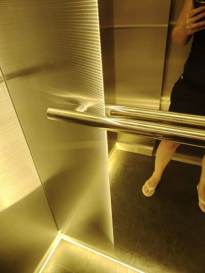 An indoor shot shows a person's legs and feet in white flip-flops standing on a dark elevator floor. The elevator walls are made of reflective, brushed gold-toned metal, with horizontal ridges visible on the left wall. Two horizontal, shiny chrome handrails are mounted on the right wall. The lighting is warm and reflects off the metallic surfaces. The person is wearing a black outfit. The scene is an elevator, likely in an urban setting, but there is no specific information to confirm it is in Chaiky, Ukraine.