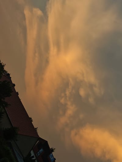 The image displays a tilted view of a street scene in Bautzen, Germany, under a dramatic sky filled with orange and gray clouds. A building with a red tiled roof is prominent on the left side of the frame, angled upwards. Adjacent to it, another section of a building is visible, featuring windows and brickwork. Green foliage, possibly trees or bushes, is present along the roofline and beside the buildings, appearing dark against the sky. The sky itself is characterized by swirling clouds with distinct areas of bright orange illumination, suggesting sunset or sunrise. The overall impression is one of natural beauty combined with urban architecture.