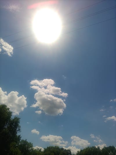 In Petrivka, Ukraine, the image captures a bright, sunny day with a clear blue sky. The sun, a brilliant white orb with rays emanating outwards, is prominently positioned in the upper left quadrant, casting a strong glare. Three thin, dark power lines stretch horizontally across the sky, intersecting the sun's glare. Below, fluffy white cumulus clouds are scattered across the blue expanse. The lower portion of the image features a dense line of lush green trees, suggesting the presence of a forest or park area. The overall scene is one of natural beauty under a bright sky, possibly indicating a warm, pleasant day.