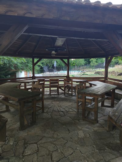 The image depicts an outdoor wooden gazebo with a stone floor, furnished with several rustic wooden picnic tables and benches. The gazebo has a multi-tiered, dark wood roof with decorative trim. Inside the gazebo, the tables are arranged in a circular pattern, with a distinct starburst design on the stone floor beneath them. Through the open sides of the gazebo, a view of the surrounding natural environment is visible, including lush green trees and vegetation. In the background, beyond a wooden fence, a swimming pool area is visible, complete with lounge chairs and a table under an umbrella. Further in the distance, there are more trees and a stone wall. The overall scene suggests a recreational area, possibly in a rural setting. The lighting indicates it is daytime, likely afternoon, with overcast or diffused sunlight given the absence of strong shadows. There is no visible text in the image. The location context is Gramatikovo, Bulgaria.