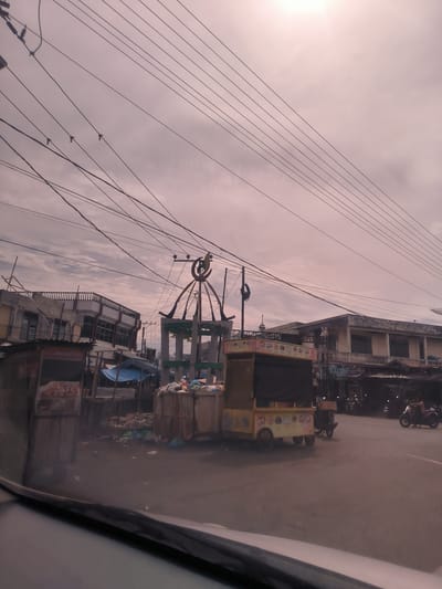 This image captures a street scene in Teungoh Baro, Indonesia, during an overcast day. The sky is a prominent feature, covered by a light pinkish-grey layer of clouds. Numerous dark power lines crisscross the upper portion of the frame, some with small insulators or bird-like objects attached.
In the midground, a bustling urban environment unfolds. Dominating the center is a decorative, possibly monument-like structure, featuring an ornate frame and topped with a large, green crescent moon and star symbol, suggestive of an Islamic context, perhaps a mosque finial or decorative archway. Around this structure and extending to the left, various makeshift stalls and market structures are visible. One prominent yellow food cart with wheels and a dark canopy sits on the right, displaying images of food products. To its left, another stall, partially obscured, shows text that includes "ZIBU KFC" and "KFC," indicating a fried chicken vendor. Piles of trash and debris are noticeable around these stalls and the central structure, hinting at a less regulated waste disposal system.
Several multi-story buildings, appearing somewhat aged with balconies and shuttered windows, line the street. One building on the left has white railings on its upper floor. On the right side of the street, a person is seen riding a motorcycle, suggesting daily transport and activity. The photograph itself is taken from inside a vehicle, with the top of a dashboard or car interior visible in the foreground at the bottom of the image. The overall scene depicts a functional, busy street with a blend of permanent structures and temporary market setups, under a muted, cloudy sky.
