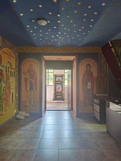 The image shows the interior of an Orthodox church in Condrița, Moldova. The ceiling is painted dark blue and adorned with small, gold star-like decorations. A central light fixture is visible on the ceiling. The walls are decorated with religious icons painted in a traditional style.
On the left wall, a large icon depicts several figures, including one labeled "Sf. Voievod Constantin Brancoveanu cu fiii". In the center of the image, an arched doorway leads to another part of the church. On either side of this doorway, within arched niches, are large icons of saints. The icon on the left depicts a bearded male saint holding a scroll. The icon on the right depicts a female saint with a veil, holding a cross and a scroll.
In the doorway, a triptych icon is visible in the distance, depicting religious figures. The floor is tiled with large, square, light-colored tiles. To the right of the central doorway, a dark wooden structure with metal accents is partially visible, possibly part of a display or altar.
The overall impression is of an ornate, sacred space with rich religious artwork. The lighting suggests it might be daytime, with some natural light entering from the doorway.