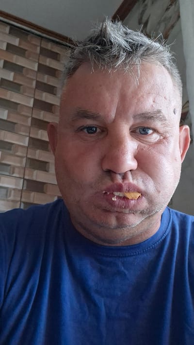 A close-up selfie shows a middle-aged man with graying, spiked hair, and blue eyes. He is wearing a blue t-shirt and has a piece of yellow food in his mouth, causing his cheeks to bulge. The background features a wall with horizontal, patterned tiles and a lighter, textured surface. There is no indication of Voinova, Moldova, in the image itself.