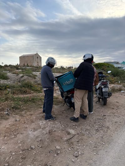 The image captures two individuals, likely delivery riders, standing with their scooters on a dirt and gravel path in a rugged, outdoor setting, consistent with the rocky landscape of Dingli, Malta. The sky above is overcast and cloudy, indicating daytime with soft, diffuse light.
In the foreground, one person wears a black puffer jacket, khaki pants, and a white helmet with a black stripe. They are standing next to a black and white scooter. Attached to the rear of this scooter is a teal blue delivery bag featuring the text "Wolt Partner" in white. The scooter's license plate, "ACR 495," is clearly visible.
To the left, another person, wearing a dark blue patterned long-sleeve shirt, dark jeans, and a grey helmet, stands next to a second scooter. This individual is turned away from the viewer, looking towards the distant scenery.
The ground is characterized by loose dirt, small stones, and sparse, dry vegetation. In the background, an old, square-shaped stone building, possibly a chapel given the small cross on its roof, sits atop a rocky hill, adding a historical and local context to the scene. The overall impression is of a pause or waiting period for the delivery drivers in a picturesque, rural Maltese landscape.
