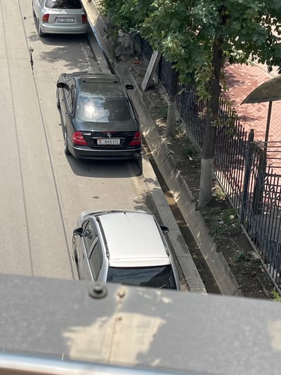 This is an aerial shot of a street in Osh City, Kyrgyzstan, featuring several parked cars. The foreground is dominated by a gray metal railing. Below the railing, a silver sedan is parked on the right side of the road, angled slightly away from the viewer. To its left, a black Mercedes-Benz sedan is parked, its license plate showing "06 644 ATU". Further up the road, a light-colored sedan is visible, with its license plate reading "284 AET". The road appears to be paved, with a concrete drainage ditch running alongside the parked cars. Trees with green foliage line the right side of the street, behind a black metal fence. A brick-patterned walkway is visible beyond the fence. The scene is illuminated by sunlight, casting shadows on the road and the cars.