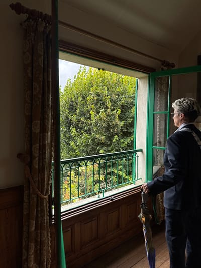 The image captures an interior view looking out through an open window in what is likely Claude Monet's house in Giverny, France. In the right foreground, an adult individual with short, grey, curly hair and glasses stands facing left, observing the exterior through the window. They wear a dark suit jacket over a lighter garment, with a diagonal striped sash across their left shoulder. Their right hand holds a multi-colored, folded umbrella that rests on the dark wooden floor.

The large, open window or French door features a green frame. Beyond it, a dense canopy of vibrant green tree foliage dominates the view. Below the foliage, a dark green metal balustrade with decorative arches is visible, suggesting a balcony. To the left of the window, a patterned curtain in beige and light brown tones, with a floral or circular motif, is tied back with a braided rope. The interior wall below the window is clad in dark wooden paneling with recessed square designs. The ceiling is light-colored. The ambient light indicates an overcast or partially cloudy sky outside.