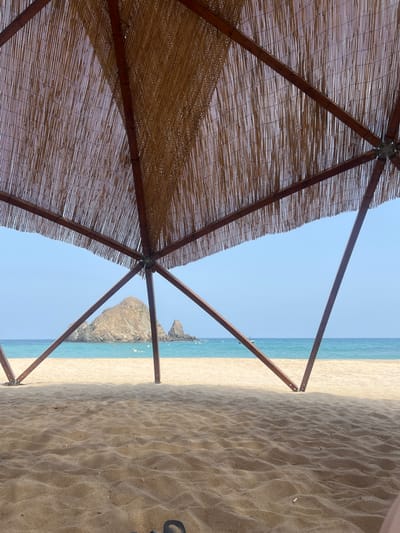 This is a low-angle, eye-level shot of a beach scene in Al Aqah, United Arab Emirates, taken from underneath a thatched roof structure. The foreground is dominated by rippled sand. Behind the sand, a strip of turquoise water meets a sandy shoreline. In the distance, a large, rocky island formation is visible in the ocean, with a smaller rocky outcrop to its right. The sky is a clear, pale blue. Wooden beams and the thatched roof of the overhead structure frame the upper portion of the image, casting shadows on the sand. A small white boat is visible on the water near the island. There are a few buoys in the water. No people are clearly visible, though the feet of a person wearing dark footwear are partially visible in the bottom right corner of the frame, suggesting the viewer is seated. The overall impression is of a serene beach setting, providing shade from the sun.