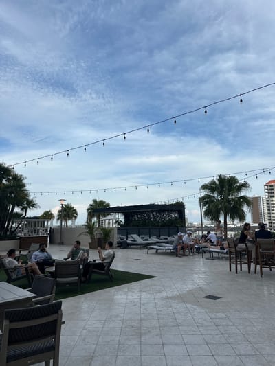 The image depicts an outdoor lounge or patio area, possibly a rooftop setting, with a view of the city skyline in Tampa, Florida, USA. The scene is captured during daylight, under a sky with scattered clouds and suspended string lights.

People are seen sitting on various chairs and benches, either alone or in small groups. They are engaged in leisurely activities like conversation, relaxation, or using electronic devices. The patio features a combination of tiled flooring and patches of artificial turf. Potted plants and palm trees add to the ambiance.

The scene includes architectural elements such as pergolas or shaded structures, which provide a break from the sun. In the background, taller buildings of the city are visible, giving context to the location. The presence of outdoor furniture and the general setting indicates a commercial or recreational space for gatherings.