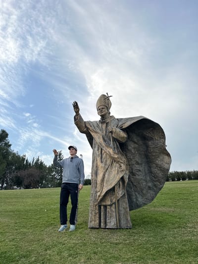 This image depicts an outdoor scene in the Galil Elyon Regional Council, Israel. A large bronze statue, approximately 1.5 to 2 times the height of a human, dominates the center-right. The statue portrays Pope John Paul II, wearing a papal mitre, cassock, and a voluminous cloak. His right arm is raised in a blessing or waving gesture, and his left hand grips a staff topped with a crucifix. To the immediate left of the statue stands a male individual, facing partially towards the camera. He is dressed in a grey hooded sweatshirt, black athletic pants with a white side stripe, and light blue athletic shoes, wearing a grey baseball cap with sunglasses resting on its brim. The man has his right arm raised, mimicking the statue's gesture. The subjects stand on a green grassy lawn. In the background to the left, a cluster of trees, including both bare deciduous and evergreen varieties, is visible. The sky above is blue with scattered white, wispy clouds. Distant vegetation lines the horizon on the right side of the image.