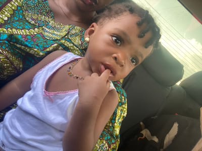 A young child lies on a brightly patterned blue and green African print fabric. She wears a white undershirt trimmed in pink, a colorful necklace, and a gold earring. Her dark hair is styled in twists. The child looks directly at the camera with her fingers in her mouth. The background shows the interior of a car, with dark gray seats and a window partially visible.
