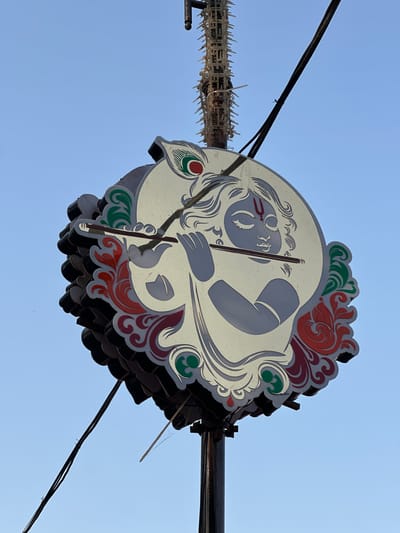 A low-angle shot shows a sign depicting Krishna in Mathura, India. The circular sign, viewed from below, features a light-colored background with a grey image of Krishna holding a flute. Red and green floral designs border the image. The sign appears to be made of multiple layers. Black electrical wires run across the blue sky in the background. A structure with pointed decorations sits near the left edge of the sign.