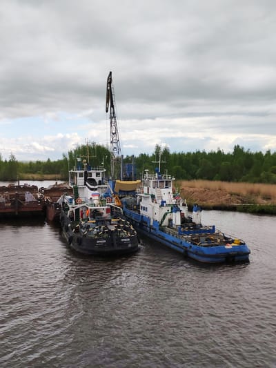 The image shows two tugboats moored in a river or waterway in the city of Ozernoe Ust'e, Russia. The tugboats, one painted black and white and the other blue and white, are docked alongside a barge or pier on the left side of the frame. A crane is visible extending upwards from one of the boats. The water is murky and dark. The background features a treeline and a cloudy sky.