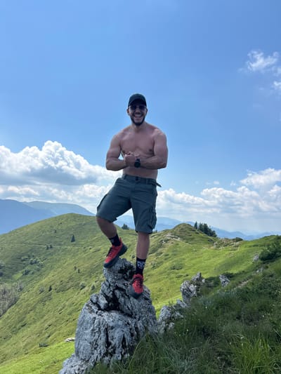 A shirtless man is standing on a rocky outcrop, with a lush green mountainside behind him. He is wearing grey cargo shorts, black sunglasses, a black baseball cap, and red hiking shoes with black socks. He is flexing his arm and giving a thumbs up to the camera, smiling. The sky is blue with fluffy white clouds. The sun appears to be high in the sky, suggesting it is midday. The location appears to be mountainous, with the man positioned on a steep slope. The greenery indicates it is likely spring or summer. The overall mood of the image is energetic and adventurous.