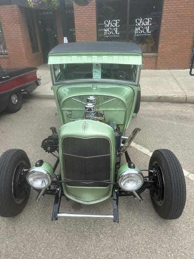 A light green 1930s-style hot rod is parked on an asphalt surface, possibly a street or parking lot, in front of a brick building. The building has large windows with "SAGE the SOL yoga & wellness" signage. A black classic car is partially visible to the left of the hot rod. The hot rod features an exposed engine with chrome detailing, a vertical grille, and prominent headlights. A small award or plaque with the text "BEST IN CATEGORY Vintage" is affixed to the windshield. The scene appears to be outdoors, with a concrete curb and sidewalk visible. The setting is the City of Leduc, Canada, indicated by address numbers "A-4726 50th AVE" and "B-4726" on the building.