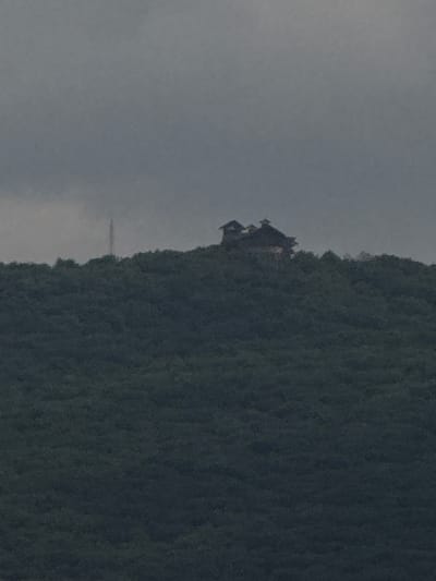The image depicts a hazy, overcast sky over a densely forested hill. Atop the hill, a dark-colored structure, possibly a house or lodge with multiple gables and dormer windows, is visible. To the left of the structure, a tall, slender tower, likely a communication tower, is present. The foliage on the hill is uniformly dark green. The overall scene suggests a remote, elevated location. There is no indication of people or specific activities. The image does not provide any geographical markers or details that would allow for definitive placement within the city of Sis, Azerbaijan.
