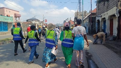 This image depicts an urban street scene in Gisenyi, Rwanda, featuring an organized clean-up activity under a clear, bright sky. Approximately fifteen individuals are visible, with the foreground dominated by five people walking away from the camera on an asphalt road and adjacent paved sidewalk.

**People:** The five individuals in the immediate foreground and midground are wearing distinctive fluorescent yellow-green and blue reflective vests, indicating participation in a clean-up operation. Most wear white protective gloves, and some carry long sticks, possibly for picking up debris. Their attire varies, including jeans, long dresses, and t-shirts. One person on the far right, wearing a brown jacket and blue jeans, is actively bent over on the brick sidewalk, appearing to collect trash into a white bag. Other individuals in similar vests are further down the street, also walking.

**Environment and Structures:** The street comprises a dark asphalt road and a lighter, reddish-brown brick-paved sidewalk on the right. Utility poles with numerous overhead power lines crisscross the scene. Buildings line both sides of the street. On the right, structures appear older, featuring exposed brick, some peeling paint, and roll-down metal shutters over doorways, with colors like blue and red. Debris is visible on the sidewalk next to these buildings. On the left, buildings are a mix of lighter-colored multi-story residential structures and commercial units with teal-colored metal shutters. In the distance, a large communication tower or similar infrastructure is prominent against the sky.

**Actions and Objects:** The primary action is the group's collective movement, suggesting a community clean-up effort. Cleaning tools include the sticks carried by participants and the white trash bag utilized by the individual on the sidewalk. The reflective vests bear partially visible text, with "CLEAP" discernible on one. The scene conveys an active, organized effort to maintain street cleanliness.