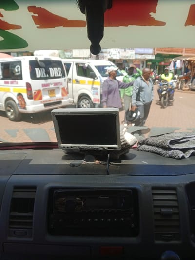 View from inside a vehicle. A Pioneer car radio and a small monitor sit on the dashboard. Outside, two white vans are visible, one decorated with "DR. DRE" and an image of a man. People are standing on the street, including one person in a green shirt and another wearing a yellow vest on a motorcycle. Buildings line the street in the background.
