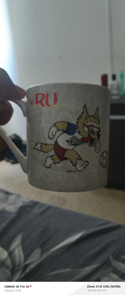 A person holds a white mug with a cartoon mascot on it. The mascot is a wolf wearing a white shirt with "RUSSIA 2018" printed on it, red shorts, and red goggles. The mug also features the letters "RU" in red. The mug is the main focus of the image, and the background is blurred. The photo was taken with a CAMON 30 Pro 5G at 23mm, f/1.8, 1/25s, ISO1554, on February 1, 2026, at 14:35, at the coordinates 51°29'4"N 128°10'1"E.