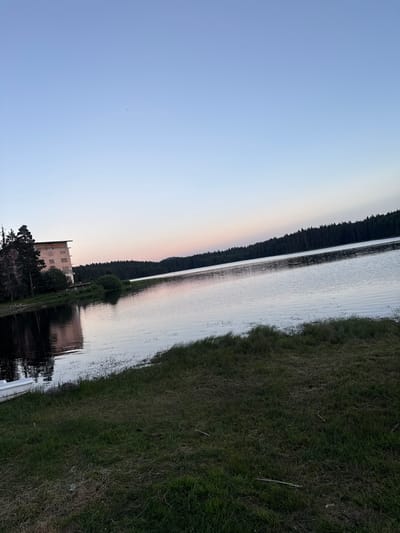 The image depicts a serene natural landscape at twilight. The scene is dominated by a large body of water, likely a lake or reservoir, reflecting the soft colors of the sky. To the left of the water, a dense, dark forest lines the shore, creating a sharp contrast with the lighter hues of the sky and water. A faint pinkish-orange glow is visible near the horizon, indicating the sun has recently set or is about to rise.
On the right side of the water, a grassy embankment slopes upwards, covered in lush green grass. In the foreground, towards the bottom left, a partially visible building with a faded, textured facade stands near the water's edge. It appears to be an older structure, possibly abandoned or under renovation, with a few trees growing around it. A portion of a white structure, possibly a boat or a pier, is visible at the very bottom edge of the image, extending into the water.
The sky is a clear, pale blue, gradually transitioning to a lighter shade towards the right edge of the frame. There are a few very small dark specks in the sky, which could be birds or debris. The overall mood of the image is peaceful and tranquil, characteristic of dusk in a natural setting. No people or specific activities are evident, suggesting a quiet moment captured in time. The location is identified as Sarnitsa, Bulgaria.