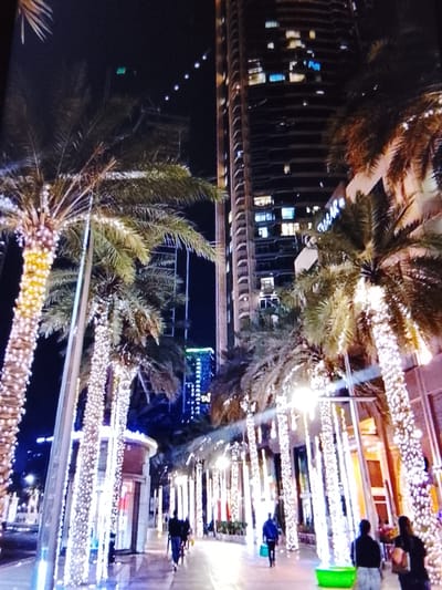 The image depicts a nighttime scene in an urban environment, characterized by illuminated palm trees and tall buildings. The palm trees are extensively decorated with strings of white lights, creating a festive atmosphere along a walkway. Several people are visible on the walkway, walking in different directions. In the background, a large, modern building with many lit windows dominates the skyline. The overall impression is that of a well-lit, possibly commercial or entertainment district at night. The specific location of Safonovo, Russia is not ascertainable from the visual information provided in the image.