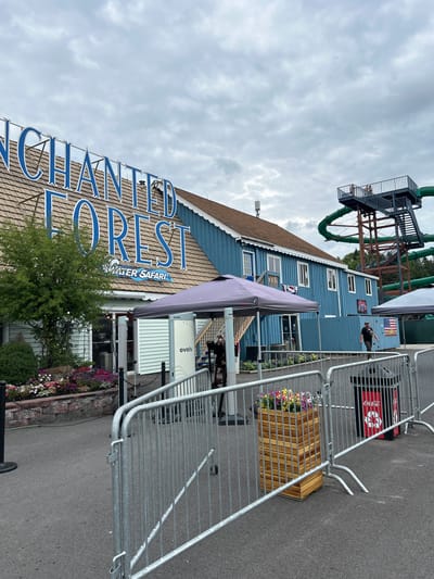 An outdoor scene at Enchanted Forest Water Safari in Old Forge, New York, features a blue building with "ENCHANTED FOREST WATER SAFARI" in large blue letters. A large green water slide structure rises behind the building. In the foreground, metal crowd control barriers line a paved area. A man in a black t-shirt and baseball cap walks past a trash can labeled "Coca-Cola" and a wooden planter filled with colorful flowers. A purple tent stands near the entrance of the building, with another tent visible further to the right. The sky is overcast.