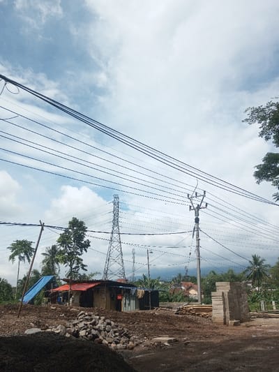 A daytime outdoor shot depicts a rural landscape dominated by power lines stretching across a cloudy sky. Below, a scene of apparent construction includes piles of gravel, lumber, and concrete blocks. A makeshift dwelling with a blue tarp and red roof is visible. An electrical tower stands in the mid-ground. Vegetation, including trees and coconut palms, provides a backdrop, suggesting a tropical or subtropical environment. A mountain range is slightly visible in the background.