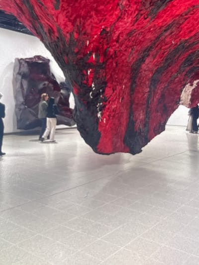 The image depicts an indoor exhibition space, possibly an art gallery, with a large, abstract sculpture dominating the foreground. The sculpture is characterized by its textured surface and bold red and black coloration, suggesting organic or raw materials. In the background, two individuals are observing the artwork. One is wearing a green jacket and light-colored pants, while the other is in a dark hoodie and light pants. Further in the background, another figure in a dark dress stands near a dark, boulder-like structure. The floor is a light-colored, polished surface marked with a grid pattern. The scene is illuminated by artificial lighting, casting soft shadows. The location in Stepantsminda, Georgia is not evident from the visual information provided in the image.