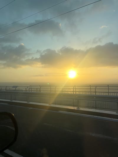The image depicts a sunrise or sunset over the ocean, observed from the Sea Cliff Bridge, near Stanwell Park, Australia. A brilliant yellow-white sun is positioned centrally on the horizon, casting intense light and lens flares across the scene. The sky transitions from pale blue at the top to luminous yellow-orange near the horizon, containing scattered grey clouds. Three dark, thin overhead lines diagonally cross the upper left sky. Below the horizon, a calm body of water reflects the sun's light. The bridge's dark asphalt surface occupies the foreground, displaying faint white lane markings. Grey metal railings delineate the bridge deck. A dark vehicle side-view mirror, partially visible in the lower left, indicates the image was captured from inside a moving vehicle.