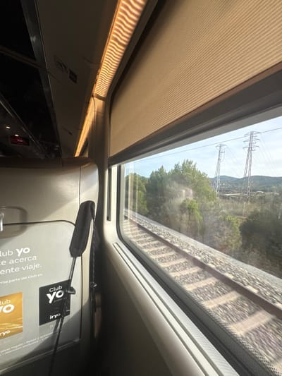 The image captures a view from inside a modern passenger train car, looking out a window towards the right. Internally, a light-colored seat or partition is visible on the left, from which a black fabric strap is suspended. Promotional stickers for "Club Yo" and "iryo" are affixed to this surface. Above the window, a beige pleated roller blind is partially deployed. A linear light fixture with a distinctive wavy pattern of illumination runs along the ceiling.

Externally, through the window, a single railway track with ballast is visible in the immediate foreground, running parallel to the train's direction. Beyond the track, a landscape of dense green trees and vegetation extends into the mid-ground. In the distance, two prominent metallic lattice-structure electricity transmission towers stand against a bright sky. Faint hills and what appears to be a distant settlement are discernible on the horizon. The scene is situated in or near Sant Esteve Sesrovires, Spain.