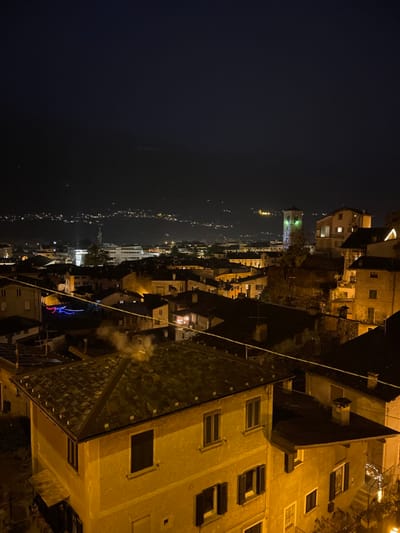 The image, when properly oriented, presents a nighttime panoramic view of a town or city, likely located in a valley or along a body of water, consistent with the location context of Sondrio, Italy. The sky above is dark, indicating a late hour, with a faint glow visible on the horizon suggesting distant light pollution or the remnants of twilight.
In the far distance, numerous small lights stretch horizontally, outlining the contours of what appears to be a long body of water or a distant winding road, flanked by illuminated settlements. Moving closer to the mid-ground, more densely packed buildings are visible, characterized by warm yellow lights emanating from windows. A prominent, taller building, possibly a church tower, stands out with its own distinct lighting. Decorative, multi-colored lights (blue, purple, red) are strung on a building or structure in this middle section, adding a festive touch.
The immediate foreground features the rooftops and upper stories of several buildings. One large building dominates the right side, showing a textured, dark-tiled roof and multiple windows, some of which are illuminated. A faint plume of white smoke or vapor is seen rising from a vent or chimney on this roof, suggesting indoor warmth or activity. Another building in the center foreground also has a visible roof. A thin wire or cable runs diagonally across the scene, from the upper right to the mid-left. No people are visible, and no discernible text appears in the image. The scene conveys a quiet, serene atmosphere of a town settled into the night.