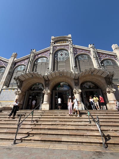 The image shows a wide-angle, low-angle shot of the exterior of the Central Market of Valencia, a historic Art Nouveau building located in Valencia, Spain. The building features an ornate facade with arched entrances, decorative tilework, and large windows. In front of the market is a wide set of stone steps leading up to the entrances.
Several people are visible on the steps and near the entrances. On the left side of the steps, a woman in black pants and a white top is standing, looking down at her phone. Near the center of the steps, a group of young adults is ascending. One person is wearing a white t-shirt and white shorts, another a white t-shirt and pink pants, and a third person a white t-shirt and beige shorts. Further into the market entrance, more people can be seen, including individuals in shorts and t-shirts. On the right side, a man in a red shirt and black shorts is standing near an entrance, and other individuals are visible further back.
The environment is sunny with a clear blue sky. The architecture is the dominant subject, showcasing intricate details and the grandeur of the market building. The presence of people suggests it is an active public space. The specific location identified is the Central Market of Valencia, Spain, which is in the city of Valencia, not Alboraia/Alboraya, which is a municipality adjacent to Valencia.