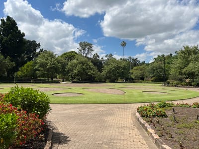 This image captures an outdoor scene, likely a park or botanical garden, presented with a horizontal orientation. The setting appears to be a sunny day, under a blue sky dotted with scattered white cumulus clouds.

In the foreground and mid-ground, a light-colored paved pathway, possibly made of pavers, curves through the landscape. Alongside this path are well-maintained garden beds, featuring lush green foliage and bursts of small red flowers.

Adjacent to the garden beds is a manicured grassy area, resembling a mini-golf course or a practice putting green. This green space includes several circular patches of bare earth or sand, and at least two small yellow flags are visible, indicating potential golf holes.

Beyond the green, a dense line of trees forms the mid-ground, showcasing a variety of tree types, including some tall, slender, palm-like trees and others with full, rounded canopies. A prominent tall, slender pole or tree trunk stands out against the sky on the left side of the frame.

No specific activities or people are visible, but the well-kept grounds and recreational features suggest a leisure environment. The bright daylight and verdant scenery indicate pleasant weather, consistent with a warm climate such as Brisbane City, Australia. There is no discernible text within the image.
