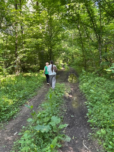 Two people are embracing on a muddy path in a lush green forest. The person on the left, with long dark hair, is wearing a white sweatshirt and grey sweatpants. They are holding the other person from behind. The person on the right is wearing a green t-shirt and black pants, and their face is not visible. The path is wet and appears to have puddles, suggesting recent rain or a damp environment. The forest is dense with tall trees and thick undergrowth, creating a secluded and natural setting. Sunlight filters through the leaves, casting dappled shadows on the path and vegetation. The overall mood appears intimate and peaceful. There is no visible text in the image. The scene likely takes place during the daytime, given the natural light. The location context provided indicates the setting is in the Hrozaŭski Sel'ski Savet, Belarus.
