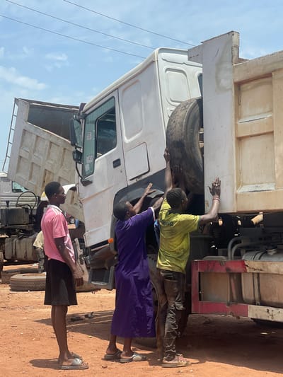 The image shows three people working on a large, white dump truck. One person, wearing a yellow shirt and dark pants, is reaching up into the engine compartment or undercarriage near a tire. Another person, wearing a purple robe, is also reaching up and appears to be assisting. A third person, wearing a pink shirt and dark shorts, stands slightly back, seemingly observing or preparing to assist. The truck is parked on a dirt or gravel surface. The sky is visible in the background and looks clear. Based on the prompt it can be assumed that the location is Kwali, Nigeria.