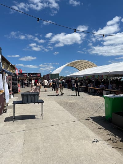 The image shows a large, outdoor gathering space with numerous people present. The location is a public space at coordinates -27.44, 153.073, which corresponds to a location near the Brisbane Street precinct in Hamilton, Australia. The scene is set under a clear, blue sky with scattered white clouds. A white canopy tent covers part of the space. There are tables with chairs for seating. A line of street food stalls with various signage is to the left. A large, black plastic storage bin is on a folding table, and a green trash can is on the right side of the image. The people are walking, sitting, standing, and interacting with each other. The event appears to be a casual gathering of people enjoying food and drinks.