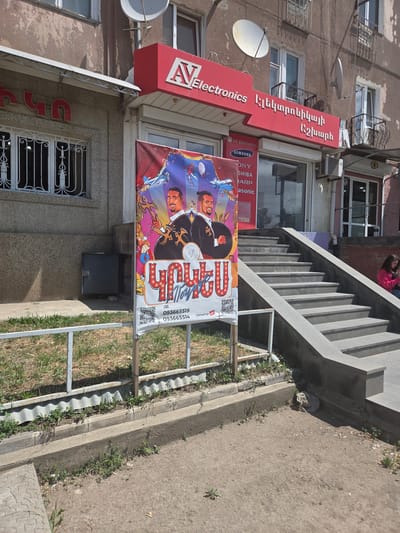 This media file depicts a street view in Gyumri-Akhuryan, Armenia. The scene is dominated by the facade of a building with an "AV Electronics" sign in red, prominently displaying the brand name in English and Armenian. Adjacent to the electronics store, a colorful banner advertises a "Circus" show, featuring cartoonish depictions of performers, including a ringmaster with a top hat and a man with a microphone. The banner also includes text in Armenian and phone numbers.

The building appears to be an older structure, with visible satellite dishes mounted on its exterior. The weather seems to be sunny, suggesting daytime. The foreground shows a grassy area bordering a concrete sidewalk and a set of stairs leading up to the building entrance. The overall impression is of a commercial street in a moderately urban area, with a mix of retail and entertainment advertising. No specific emotions are discernible from the static image.