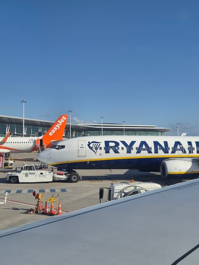 The image depicts a Ryanair aircraft parked at Francisco Sá Carneiro Airport in Moreira, Portugal, adjacent to an EasyJet aircraft. The Ryanair aircraft is white with a blue and yellow stripe along the fuselage and the Ryanair logo. The EasyJet plane is white with an orange tail fin displaying the company logo. The aircrafts are parked on a paved apron next to a modern airport terminal building with a glass facade. Ground service equipment, including tugs, baggage carts and safety cones, surround the planes. The sky is clear and blue. The angle of the photograph suggests it was taken from inside another aircraft, looking down at the other planes.